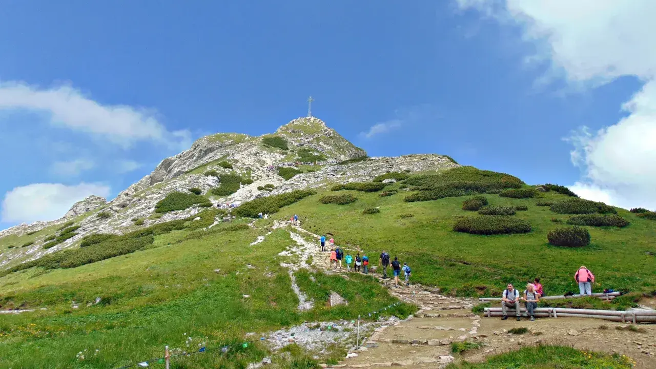 Tłum turystów wspina się kamienistym szlakiem na Giewont. Widać krzyż na szczycie.