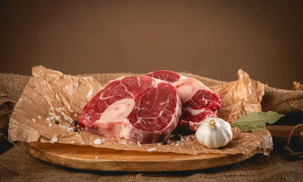 Dwa kawałki surowego ossobuco wołowego, czosnek, liść laurowy i przyprawy na desce.