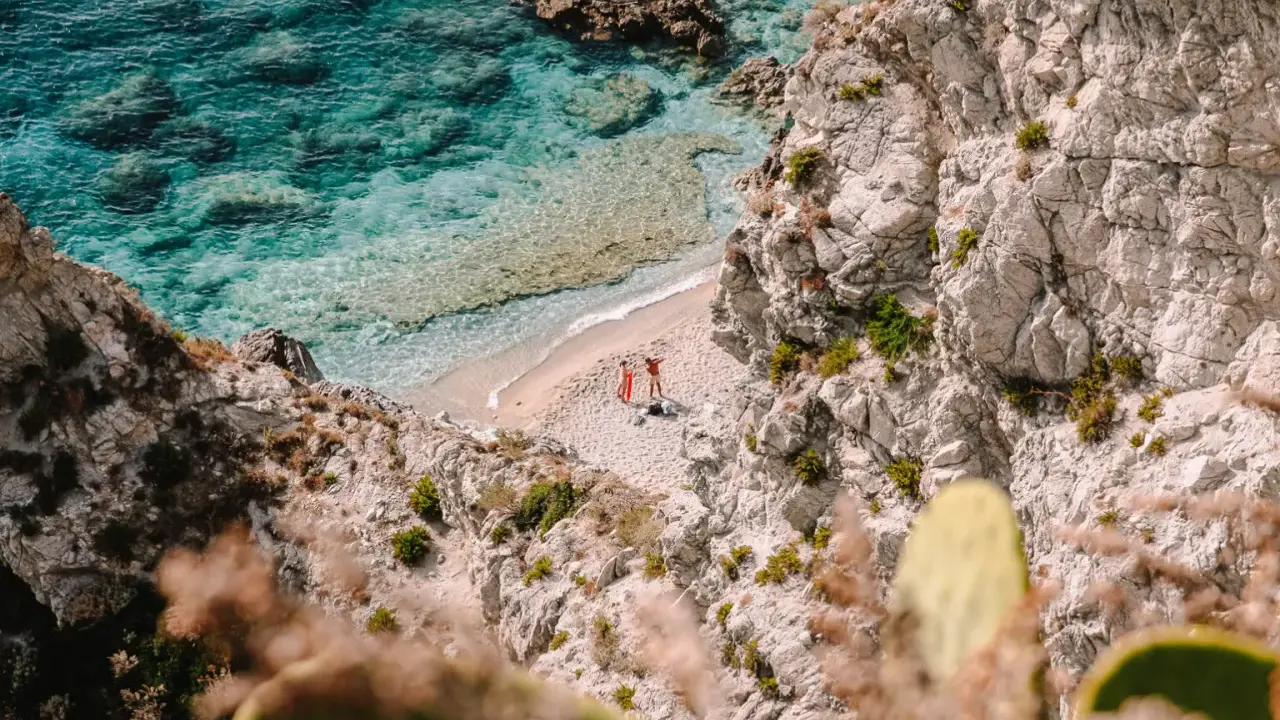 Malownicza zatoczka na Kalabrii, gdzie warto zobaczyć turkusowe morze i skaliste wybrzeże. Dwie osoby odpoczywają na piaszczystej plaży.