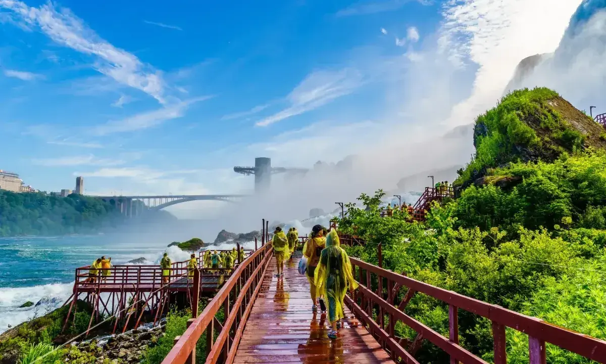 Spacerem po drewnianych kładkach przy wodospadzie Niagara, turyści w żółtych płaszczach podziwiają potęgę natury. W tle most i wieża widokowa.
