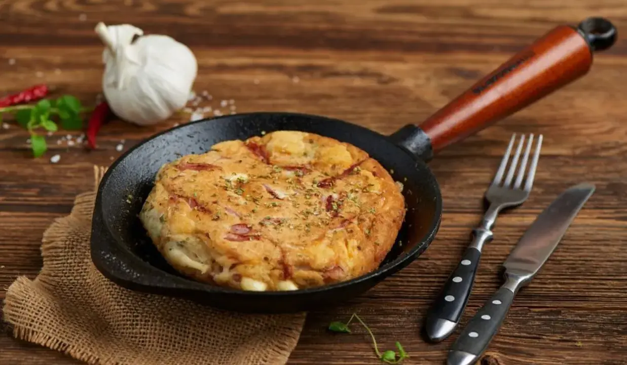 Pyszny omlet wytrawny z kawałkami wędliny, podany na patelni. Obok widelec i nóż.