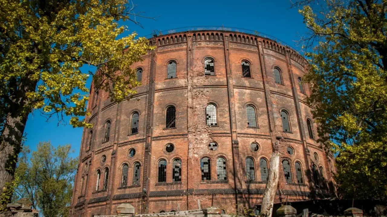 Okazały, ceglany gazometr, jeden z opuszczonych miejsc w Warszawie, otoczony jesienną zielenią.