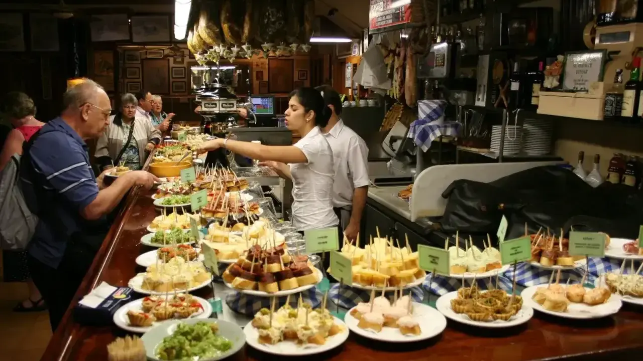 Tapas bar pełen ludzi wybierających przekąski. Na ladzie mnóstwo małych kanapeczek i innych przysmaków.