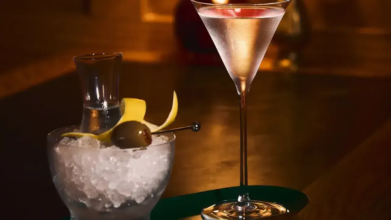 Elegancki drink z martini w kieliszku, obok miseczka z lodem, oliwką i skórką cytryny.