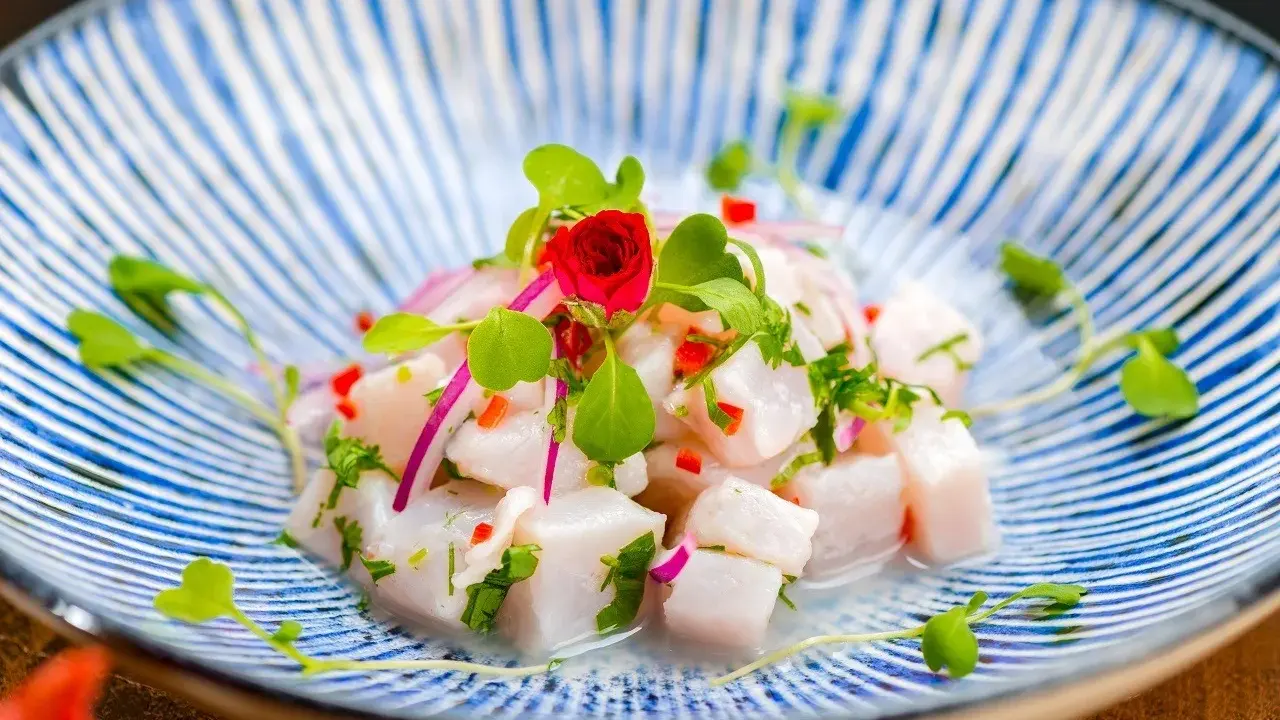 Pyszne ceviche z kawałków białej ryby, czerwonej cebuli, papryczki chili i ziół, udekorowane kiełkami i małą czerwoną różą, na niebiesko-białym talerzu.