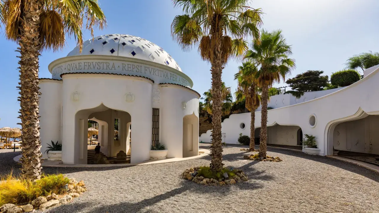 Białe budynki z kopułą i łukami, otoczone palmami i żwirem, tworzą architekturę term Kalithea.