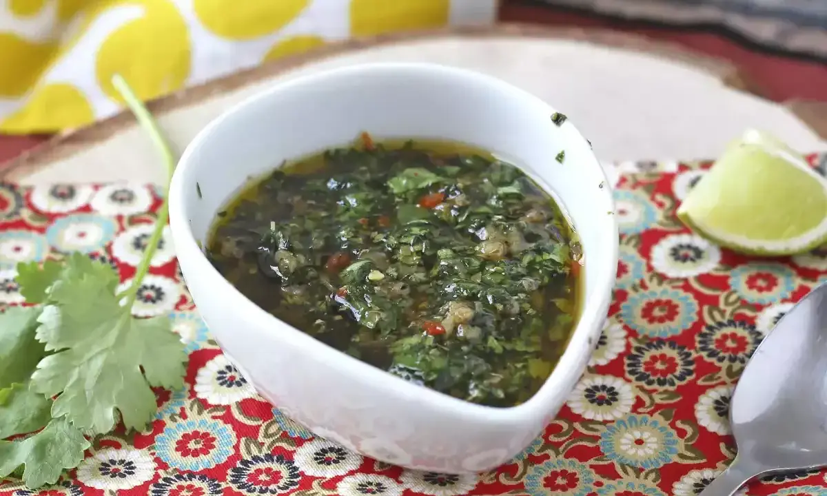 Aromaticzny sos chimichurri w białej miseczce, obok ćwiartka limonki i świeża kolendra. Idealny do grillowanych mięs.