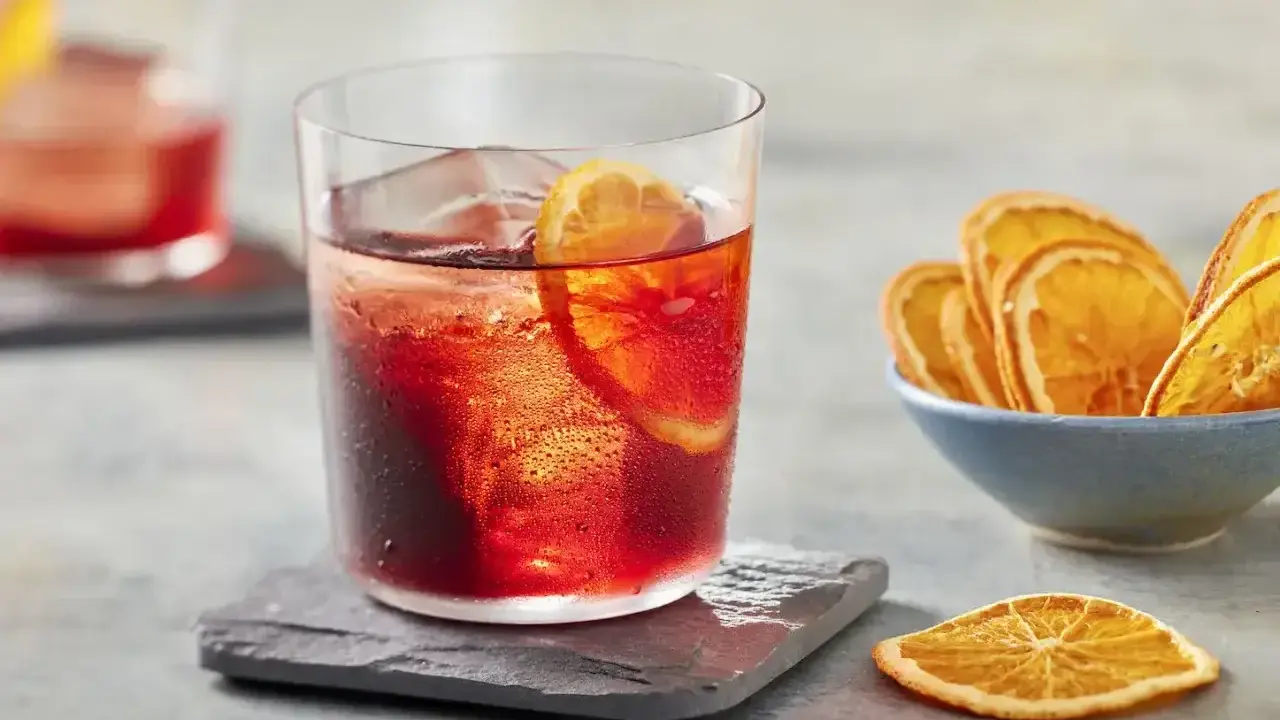Klasyczny negroni przepis: czerwony koktajl z lodem i plasterkiem pomarańczy, obok miseczka suszonych pomarańczy.