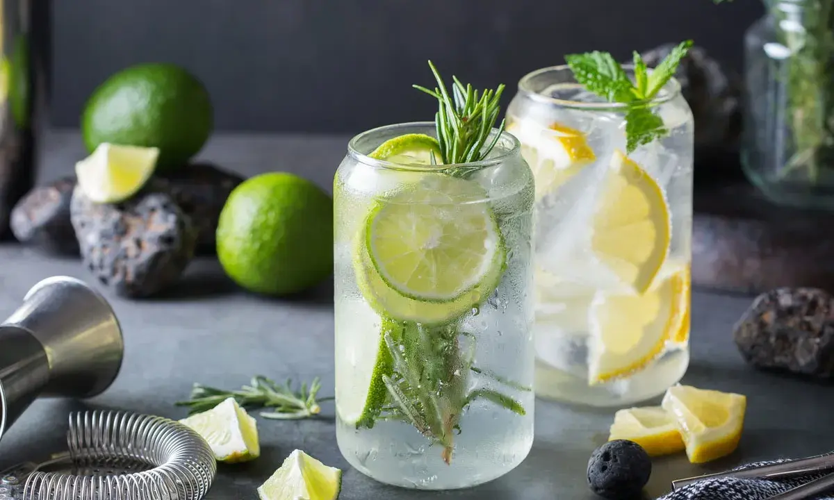 Orzeźwiający drink z ginem, limonką i rozmarynem. Obok leżą świeże limonki, cytryny i akcesoria barmańskie.