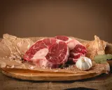 Dwa kawałki surowego ossobuco wołowego, czosnek, liść laurowy i przyprawy na desce.