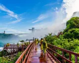Spacerem po drewnianych kładkach przy wodospadzie Niagara, turyści w żółtych płaszczach podziwiają potęgę natury. W tle most i wieża widokowa.