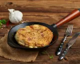 Pyszny omlet wytrawny z kawałkami wędliny, podany na patelni. Obok widelec i nóż.