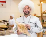Mężczyzna w tradycyjnym stroju i czapce z owczej wełny trzyma chleb gruziński. W tle widać flagę Gruzji i piekarnię.