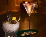 Elegancki drink z martini w kieliszku, obok miseczka z lodem, oliwką i skórką cytryny.