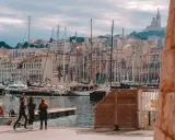 Trzy kobiety spacerują po nabrzeżu w Marsylii, w tle widać liczne żaglówki i wzgórze z bazyliką Notre-Dame de la Garde.