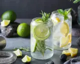 Orzeźwiający drink z ginem, limonką i rozmarynem. Obok leżą świeże limonki, cytryny i akcesoria barmańskie.