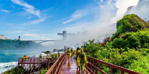 Spacerem po drewnianych kładkach przy wodospadzie Niagara, turyści w żółtych płaszczach podziwiają potęgę natury. W tle most i wieża widokowa.
