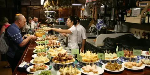Tapas bar pełen ludzi wybierających przekąski. Na ladzie mnóstwo małych kanapeczek i innych przysmaków.