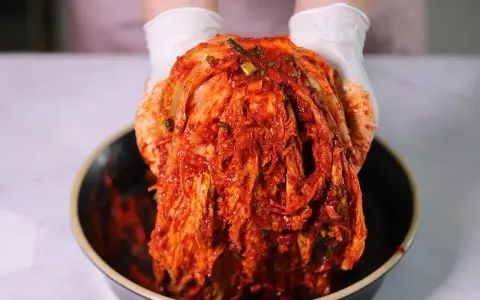 Ręce w białych rękawiczkach trzymają głowę kapusty pekińskiej pokrytą czerwoną pastą. To właśnie kimchi, tradycyjna koreańska potrawa.