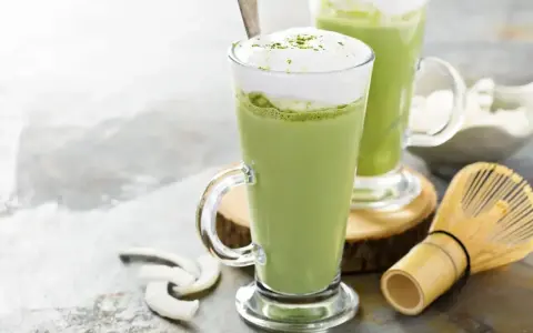 Dwa zielone napoje matcha w szklankach z pianką, obok bambusowa miotełka i wiórki kokosowe. Tak smakuje matcha.