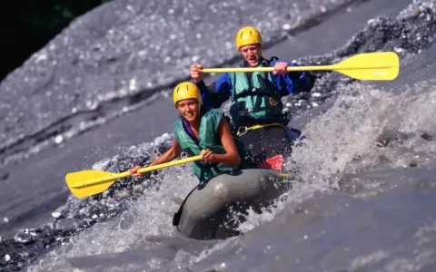 Dwóch uśmiechniętych ludzi w kaskach i kamizelkach pokonuje rwący nurt rzeki na pontonie. To właśnie rafting – emocjonująca przygoda!
