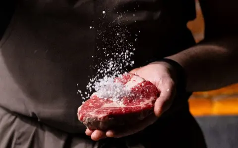 Dłoń kucharza doprawia solą soczysty kawałek mięsa, przygotowując go do grillowania. To początek drogi do idealnych rodzajów steków.
