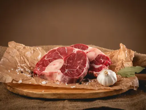 Dwa kawałki surowego ossobuco wołowego, czosnek, liść laurowy i przyprawy na desce.
