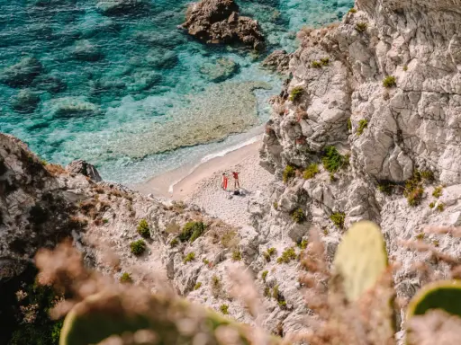 Malownicza zatoczka na Kalabrii, gdzie warto zobaczyć turkusowe morze i skaliste wybrzeże. Dwie osoby odpoczywają na piaszczystej plaży.