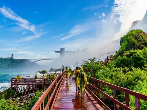 Spacerem po drewnianych kładkach przy wodospadzie Niagara, turyści w żółtych płaszczach podziwiają potęgę natury. W tle most i wieża widokowa.