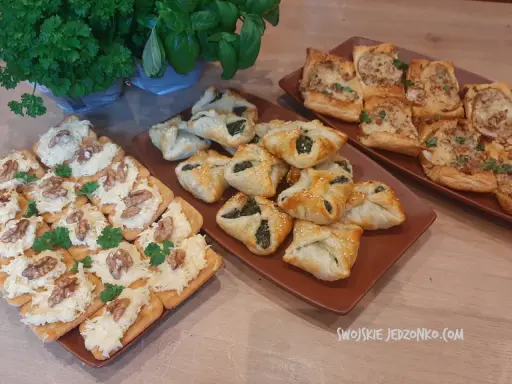 Na stole trzy rodzaje przekąsek: krakersy z pastą serową i orzechami, pieczone ciastka z nadzieniem szpinakowym i kwadratowe tartaletki z farszem. Idealna szybka przekąska na imprezę.