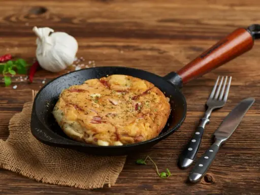 Pyszny omlet wytrawny z kawałkami wędliny, podany na patelni. Obok widelec i nóż.