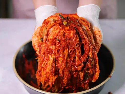 Ręce w białych rękawiczkach trzymają głowę kapusty pekińskiej pokrytą czerwoną pastą. To właśnie kimchi, tradycyjna koreańska potrawa.
