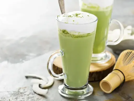 Dwa zielone napoje matcha w szklankach z pianką, obok bambusowa miotełka i wiórki kokosowe. Tak smakuje matcha.
