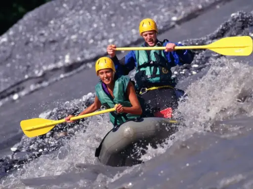 Dwóch uśmiechniętych ludzi w kaskach i kamizelkach pokonuje rwący nurt rzeki na pontonie. To właśnie rafting – emocjonująca przygoda!