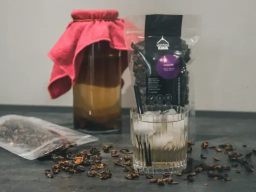 Napój z cascary, czyli co to jest kombucha z kawy, w szklance z lodem i słomką. Obok słoik z napojem i paczka z cascara.