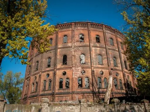 Okazały, ceglany gazometr, jeden z opuszczonych miejsc w Warszawie, otoczony jesienną zielenią.