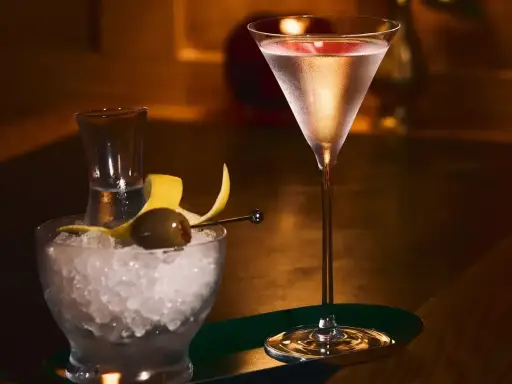 Elegancki drink z martini w kieliszku, obok miseczka z lodem, oliwką i skórką cytryny.