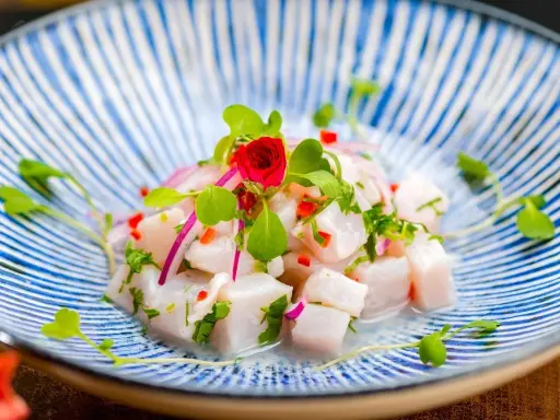 Pyszne ceviche z kawałków białej ryby, czerwonej cebuli, papryczki chili i ziół, udekorowane kiełkami i małą czerwoną różą, na niebiesko-białym talerzu.