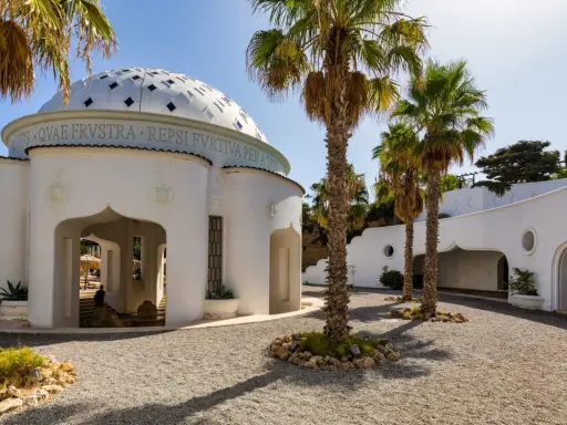 Białe budynki z kopułą i łukami, otoczone palmami i żwirem, tworzą architekturę term Kalithea.