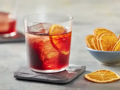 Klasyczny negroni przepis: czerwony koktajl z lodem i plasterkiem pomarańczy, obok miseczka suszonych pomarańczy.