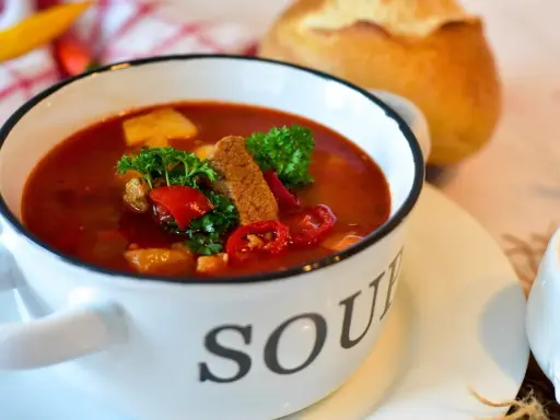 Aromatyczna zupa gulaszowa w białej misce z napisem "SOUP", ozdobiona natką pietruszki i papryką. Idealna na chłodne dni, to jedne z tych **dobrych zup domowych**.