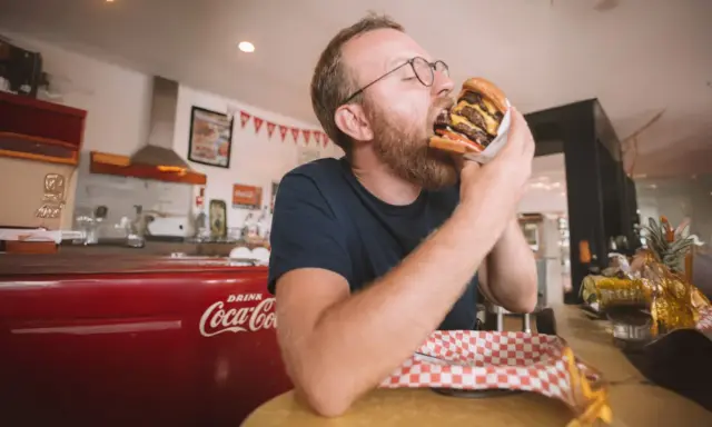 Mężczyzna z brodą i okularami zajada ogromnego burgera. W tle czerwona lodówka Coca-Coli, która może być jedną z ciekawostek o USA.