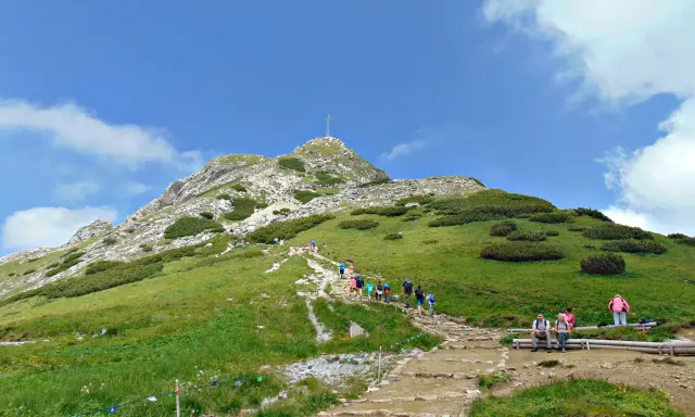 Tłum turystów wspina się kamienistym szlakiem na Giewont. Widać krzyż na szczycie.