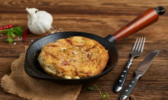 Pyszny omlet wytrawny z kawałkami wędliny, podany na patelni. Obok widelec i nóż.
