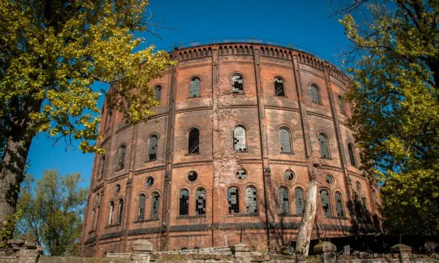 Okazały, ceglany gazometr, jeden z opuszczonych miejsc w Warszawie, otoczony jesienną zielenią.