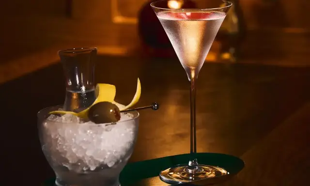 Elegancki drink z martini w kieliszku, obok miseczka z lodem, oliwką i skórką cytryny.