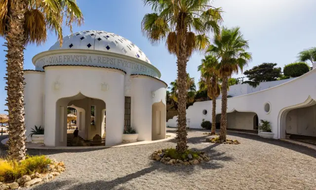 Białe budynki z kopułą i łukami, otoczone palmami i żwirem, tworzą architekturę term Kalithea.