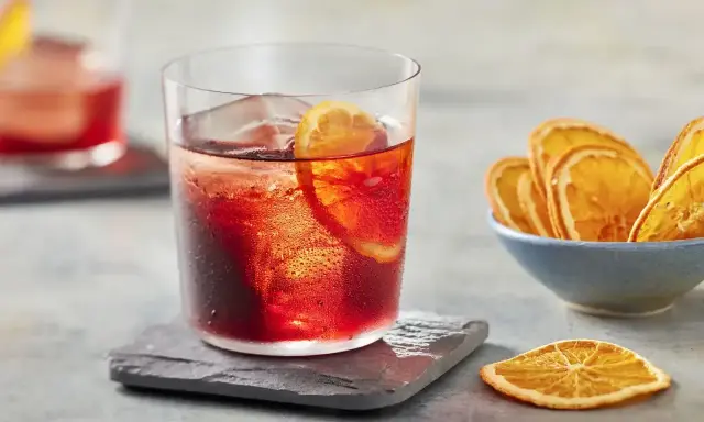 Klasyczny negroni przepis: czerwony koktajl z lodem i plasterkiem pomarańczy, obok miseczka suszonych pomarańczy.