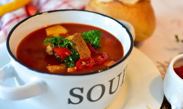 Aromatyczna zupa gulaszowa w białej misce z napisem "SOUP", ozdobiona natką pietruszki i papryką. Idealna na chłodne dni, to jedne z tych **dobrych zup domowych**.