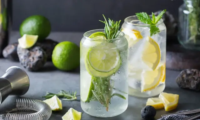 Orzeźwiający drink z ginem, limonką i rozmarynem. Obok leżą świeże limonki, cytryny i akcesoria barmańskie.